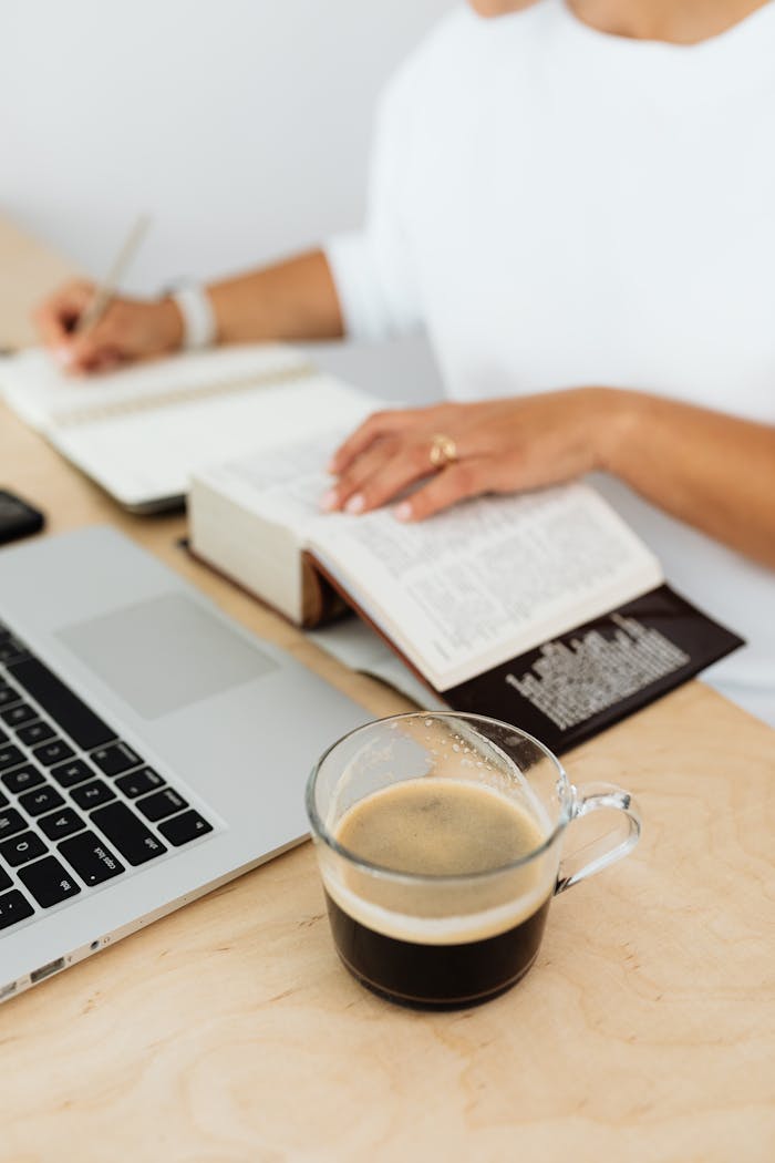 A woman writing in a notebook with coffee and laptop on a wooden desk.